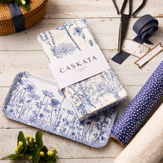 The Caskata Blooming Blues Bundle, featuring a wildflower serving tray and matching blue botanical cotton kitchen towels, is styled with ribbon, scissors, and gift wrap on a rustic white table for a charming cottagecore look.