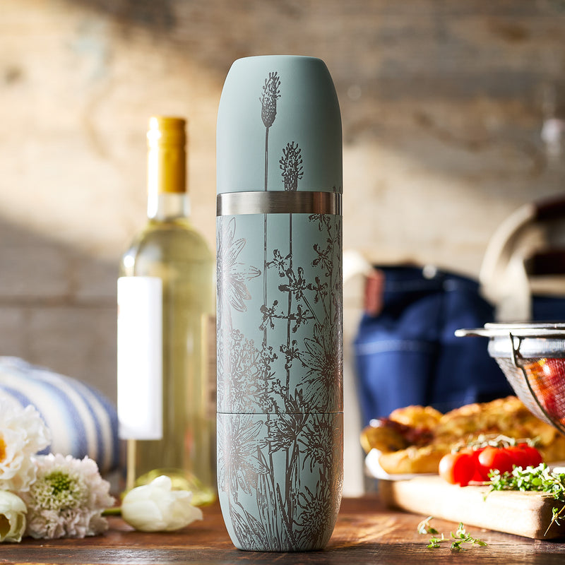 A Caskata x High Camp Light Blue Summer Parkside Flask by Caskata sits on a kitchen counter near a bottle of white wine, food, and kitchen utensils.
