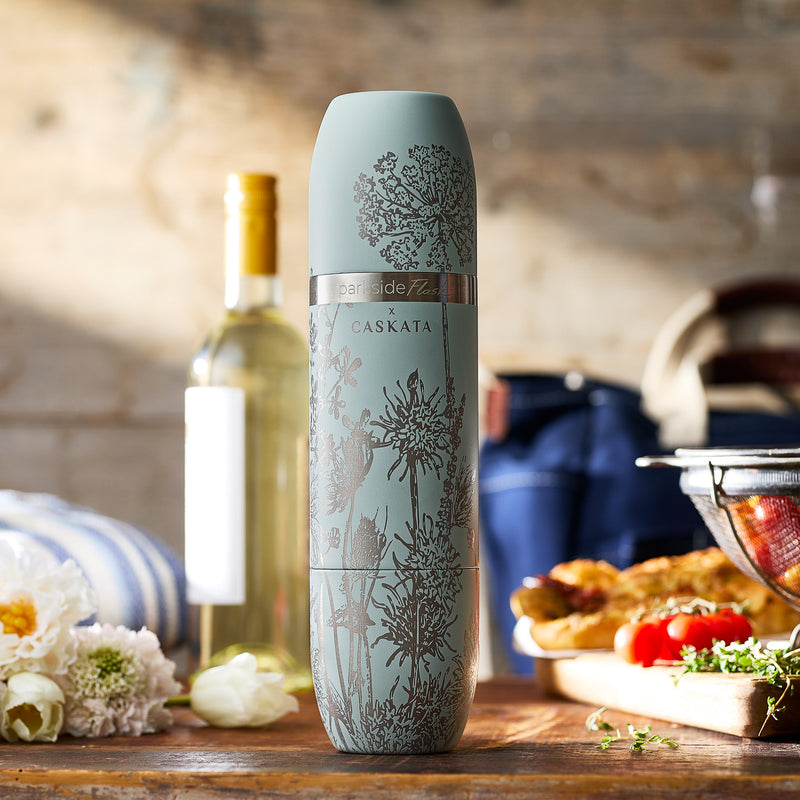 The Caskata x High Camp Light Blue Summer Parkside Flask by Caskata sits on a wooden table, surrounded by wine, food, and kitchen items in the background.