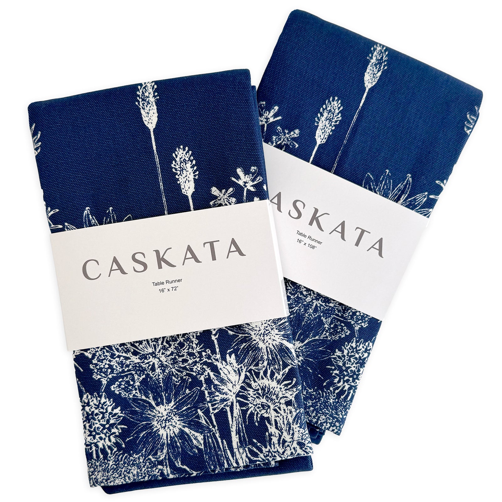 Two folded navy blue cotton table runners with white botanical prints, each wrapped in a "Caskata Summer Blues Table Runner" label—a charming addition to cottage-core or summer decor.