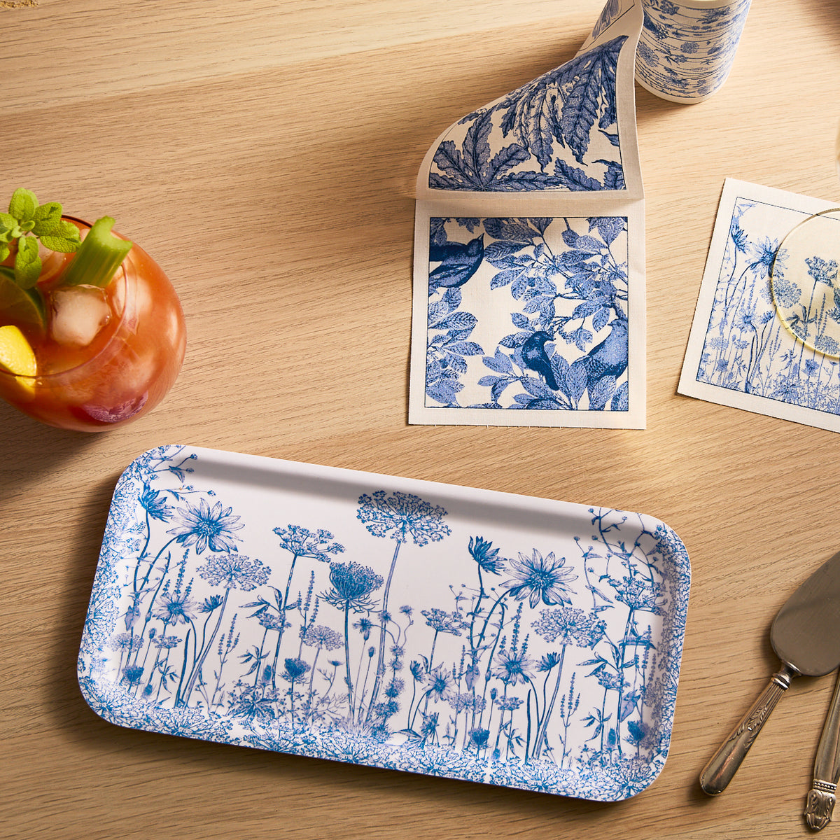 Caskata&#39;s Summer Blues Small Rectangular Birchwood Tray, featuring a blue and white floral pattern, is displayed on a botanical-themed wooden table with a fruity drink and knife.