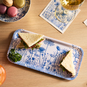 Two triangular sandwiches with lettuce and tomato are on a Summer Blues Small Rectangular Birchwood Tray by Caskata, garnished with parsley. Nearby: white wine, floral napkins, and macarons—a perfect setup to savor summer blues in an eco-friendly style.