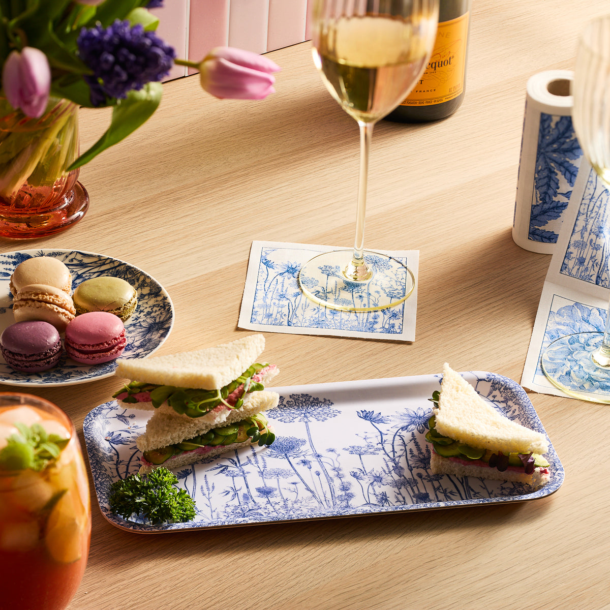 A table with triangle sandwiches on a floral plate, macarons on a Caskata Summer Blues Small Rectangular Birchwood Tray, wine glasses, and floral napkins. A flower vase and a drink complete this setting that&#39;s both charming and eco-friendly.