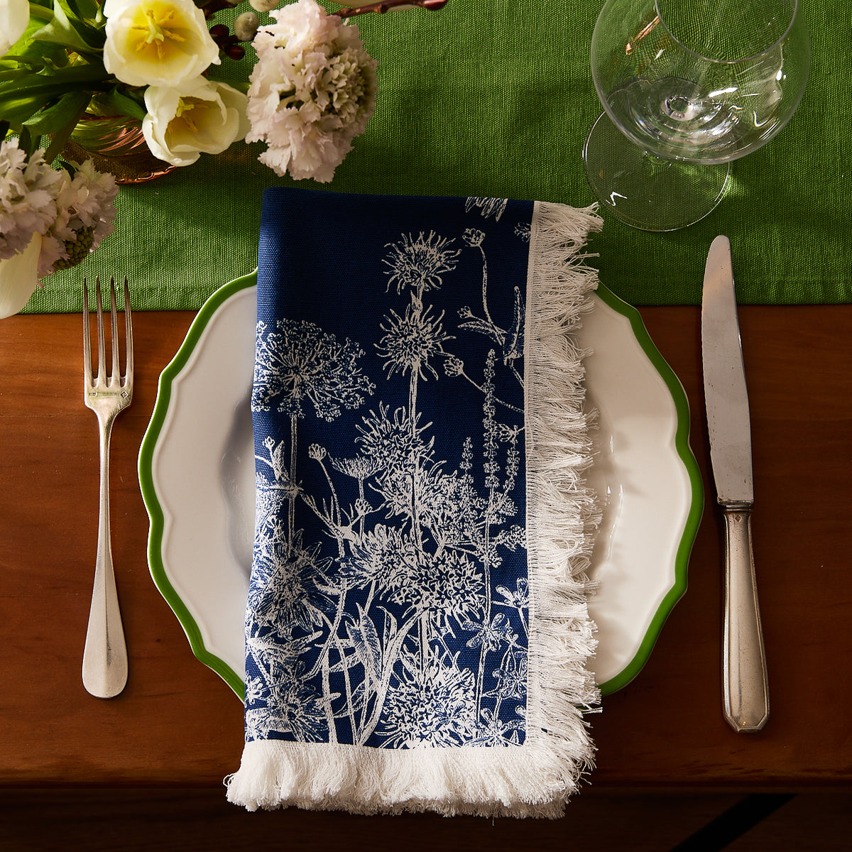 A white plate with a green rim rests on a green placemat, topped with Caskata’s Summer Blues Dinner Napkins. A fork, knife, and glass sit beside it, while flowers add a charming cottage-core touch to the table decor.