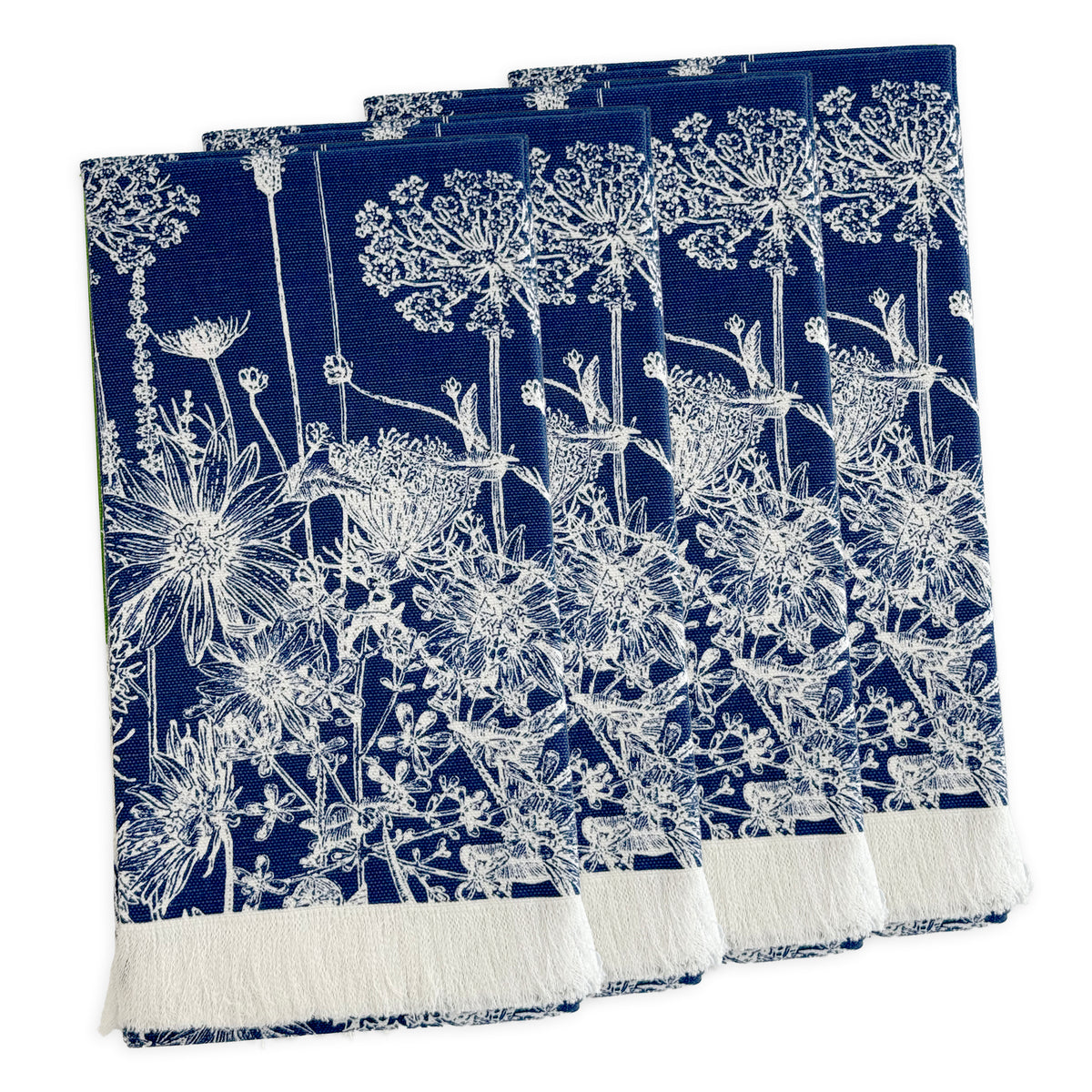 A set of four Summer Blues Dinner Napkins by Caskata in navy blue cotton with white floral patterns, inspired by New England wildflowers and finished with a white fringe on one edge, arranged in a slightly overlapping stack.