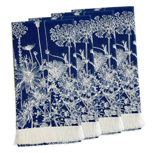 A set of four Summer Blues Dinner Napkins by Caskata in navy blue cotton with white floral patterns, inspired by New England wildflowers and finished with a white fringe on one edge, arranged in a slightly overlapping stack.