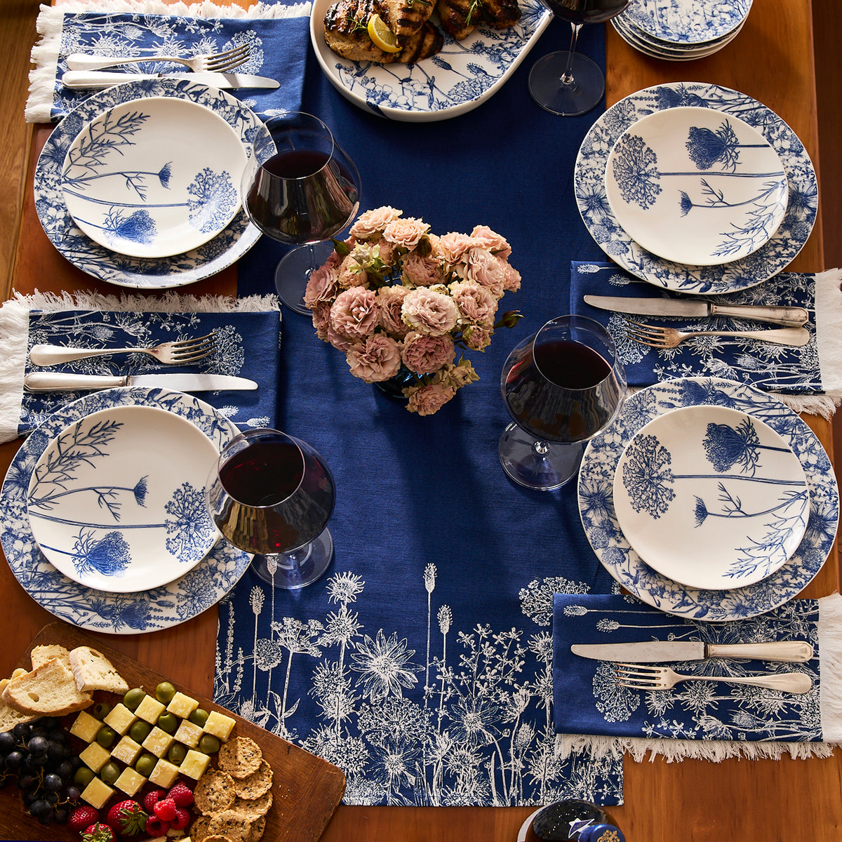 A table set for four with blue and white floral plates, blue napkins, wine glasses with red wine, a floral centerpiece, platters of grilled chicken and cheese, and the Caskata Summer Blues Table Runner—perfect cottage-core decor.