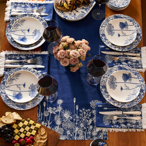 A table set for four with blue and white floral plates, blue napkins, wine glasses with red wine, a floral centerpiece, platters of grilled chicken and cheese, and the Caskata Summer Blues Table Runner—perfect cottage-core decor.