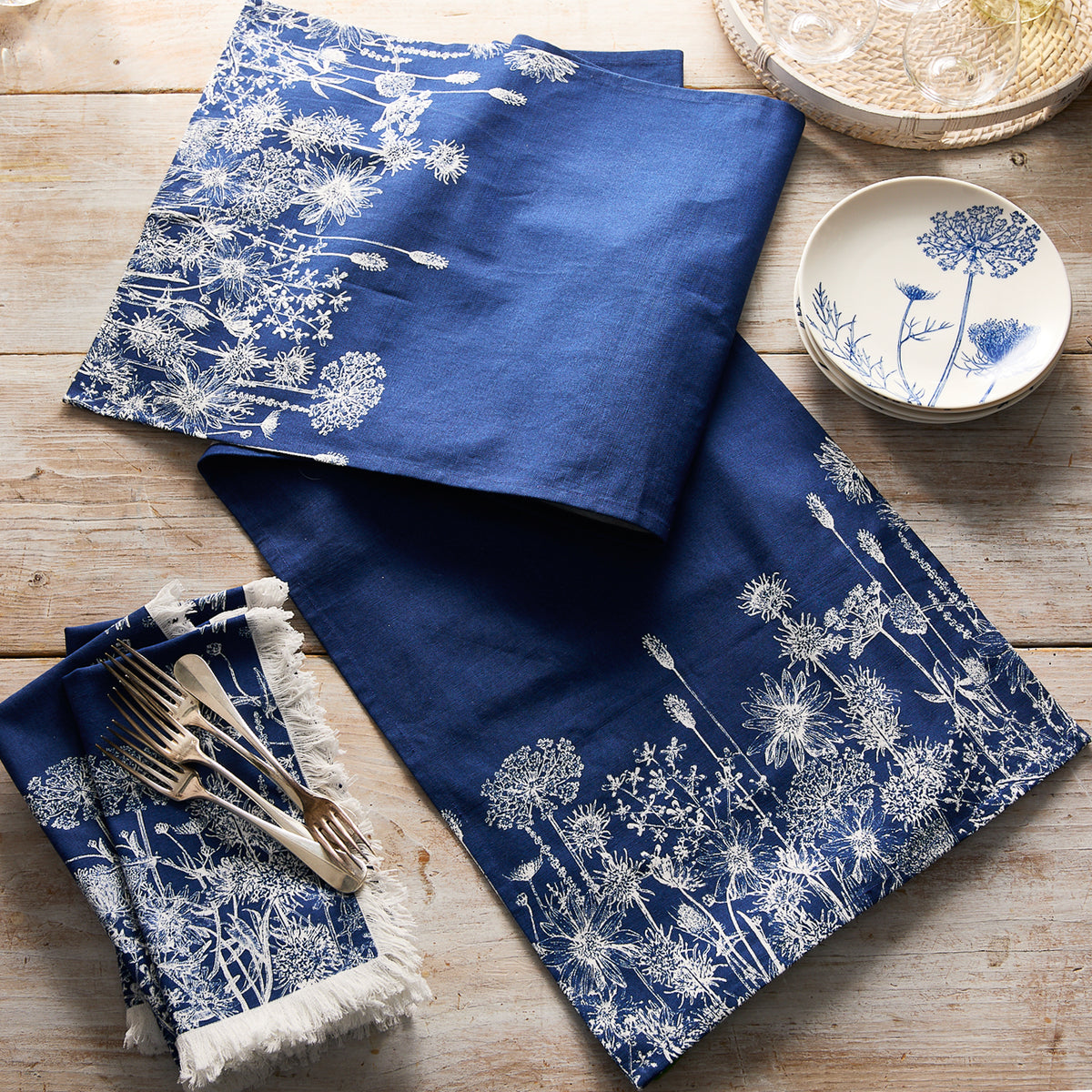 The Caskata Summer Blues Table Runner, with white floral patterns, brightens a wooden table—perfect for cottage-core decor when paired with coordinating blue and white plates and silverware.
