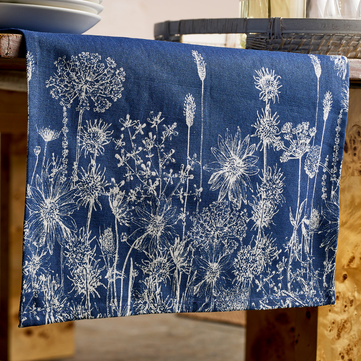 The Caskata Summer Blues Table Runner, featuring white floral and botanical patterns on blue cotton, drapes elegantly over a wooden table for a charming cottage-core touch.