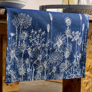 The Caskata Summer Blues Table Runner, featuring white floral and botanical patterns on blue cotton, drapes elegantly over a wooden table for a charming cottage-core touch.