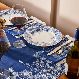 Set a charming cottage-core table with Caskata’s Summer Blues Table Runner, blue and white floral plates, silverware, wine glasses filled with red wine, and a bottle of wine for the perfect dining decor.
