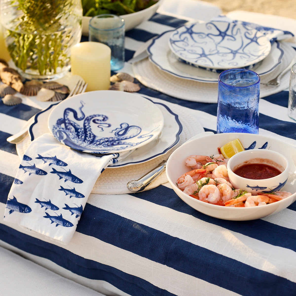 A table set with blue and white striped linens, marine-themed plates and napkins, a bowl of shrimp with sauce, candles, shells, and Caskata’s Viento Large Cobalt Tumblers (set of 6).