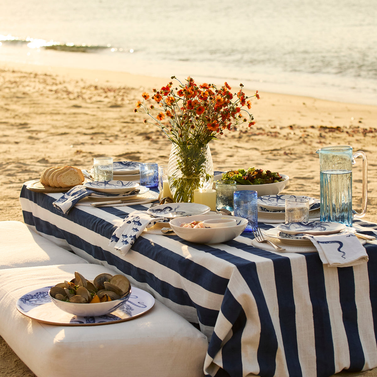 A seaside picnic features a blue and white striped tablecloth, plates of food, and Caskata Viento Large Cobalt Tumblers (Set of 6) beside orange flowers in a vase, all arranged on a low table on the sand.