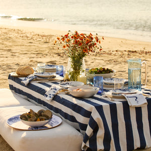 A seaside picnic features a blue and white striped tablecloth, plates of food, and Caskata Viento Large Cobalt Tumblers (Set of 6) beside orange flowers in a vase, all arranged on a low table on the sand.