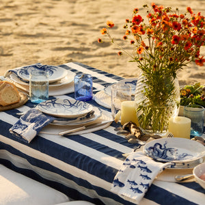 A picnic table on sand features a striped cloth, blue and white plates, Caskata Viento Medium Cobalt Tumblers (set of 6), a vase of orange flowers, candles, bread, and napkins with sea life designs.