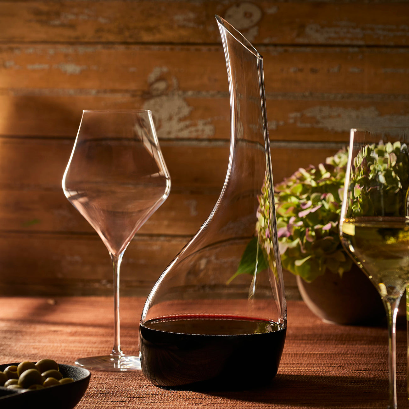 Four Caskata Terrace Red Wine Glasses stand in a row on a white background; the leftmost glass swirls red wine, while the other three remain empty.