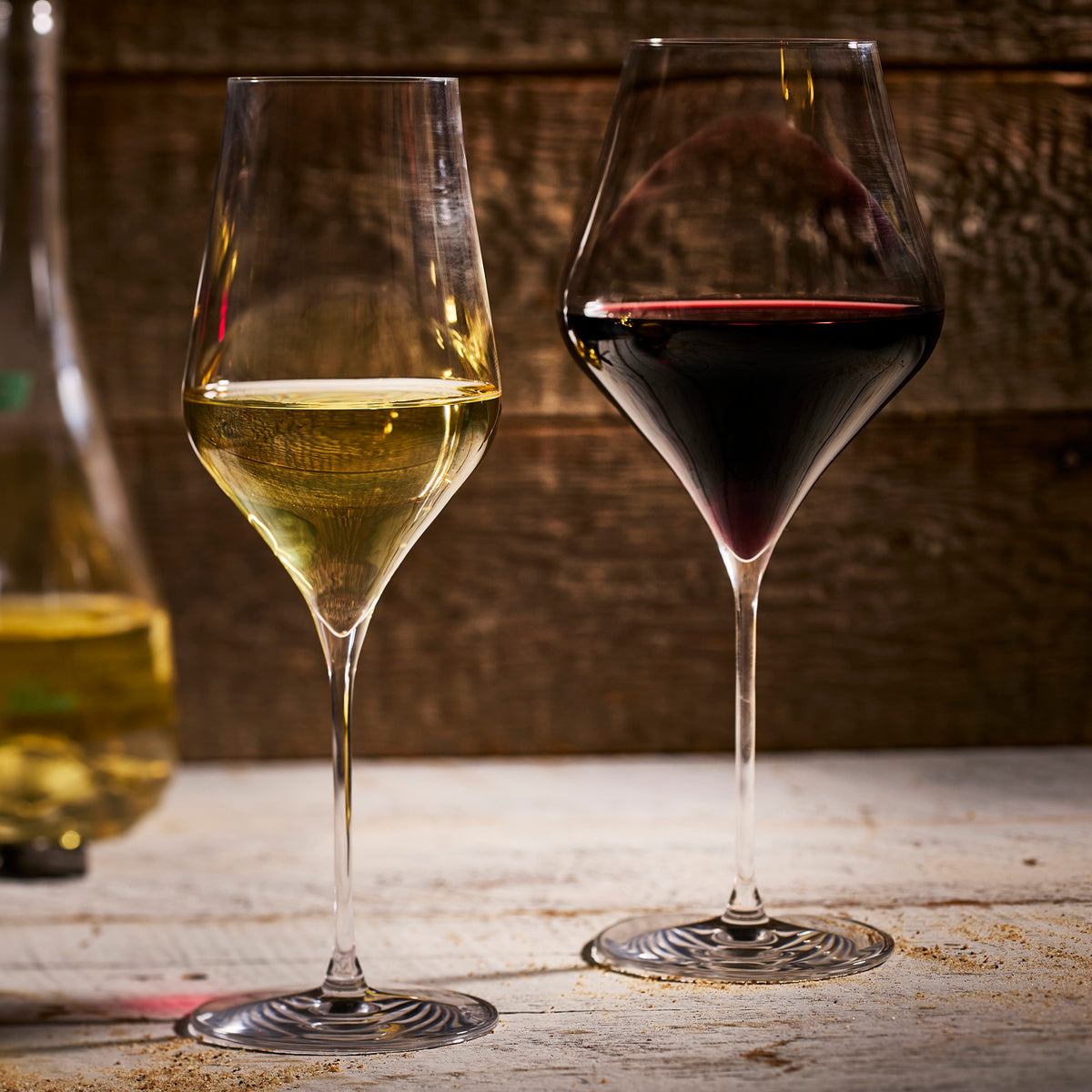 A glass of red and a glass of white wine, served in Caskata’s The Terrace Wine Lover's Starter Set lead-free crystal glasses, rest side by side on a wooden surface against a rustic wooden backdrop.