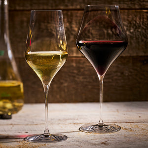 A glass of red and a glass of white wine, served in Caskata’s The Terrace Wine Lover's Starter Set lead-free crystal glasses, rest side by side on a wooden surface against a rustic wooden backdrop.