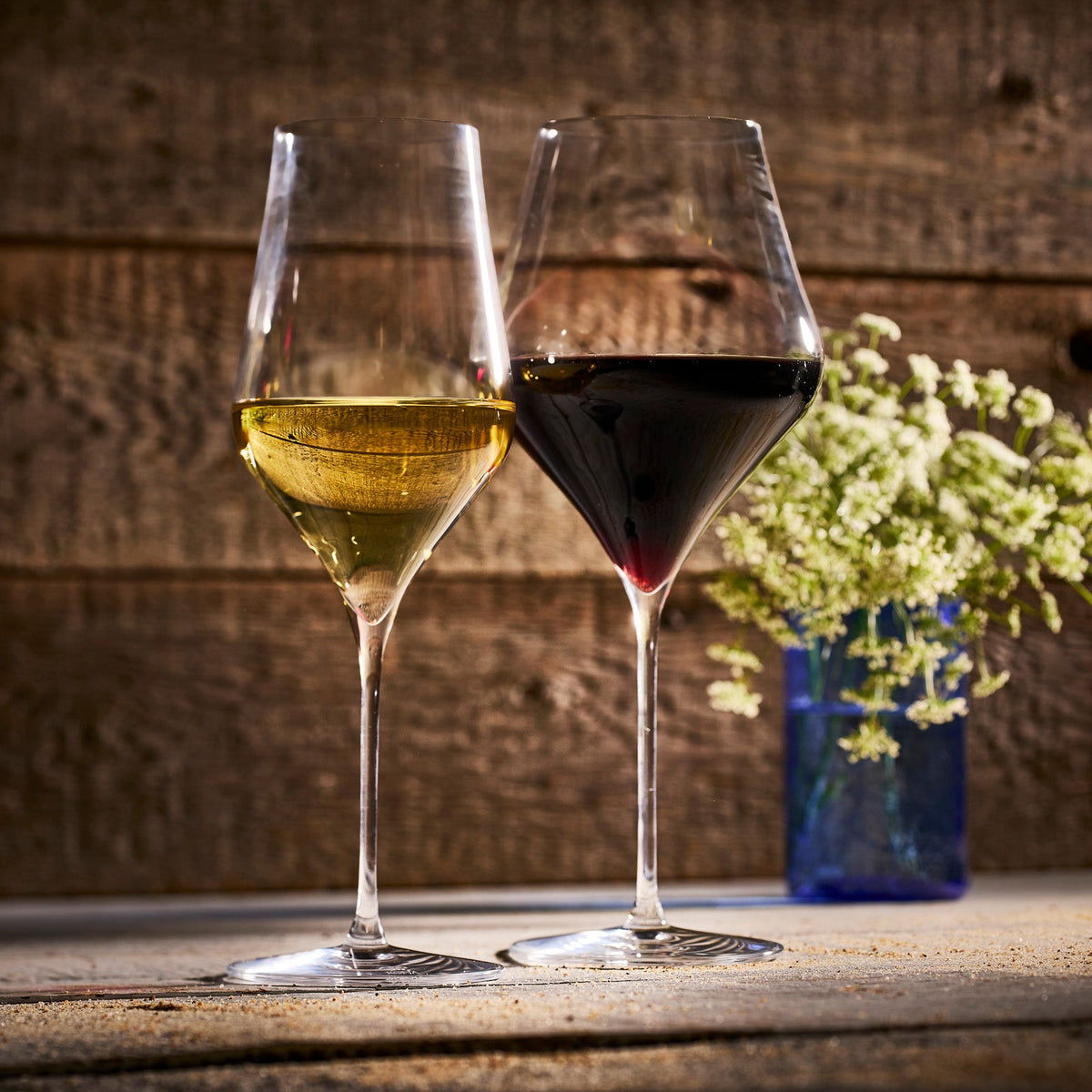 Two Terrace Red Wine Glasses by Caskata, filled with white and red wine, sit side by side on a wooden surface with a small vase of flowers in the background.