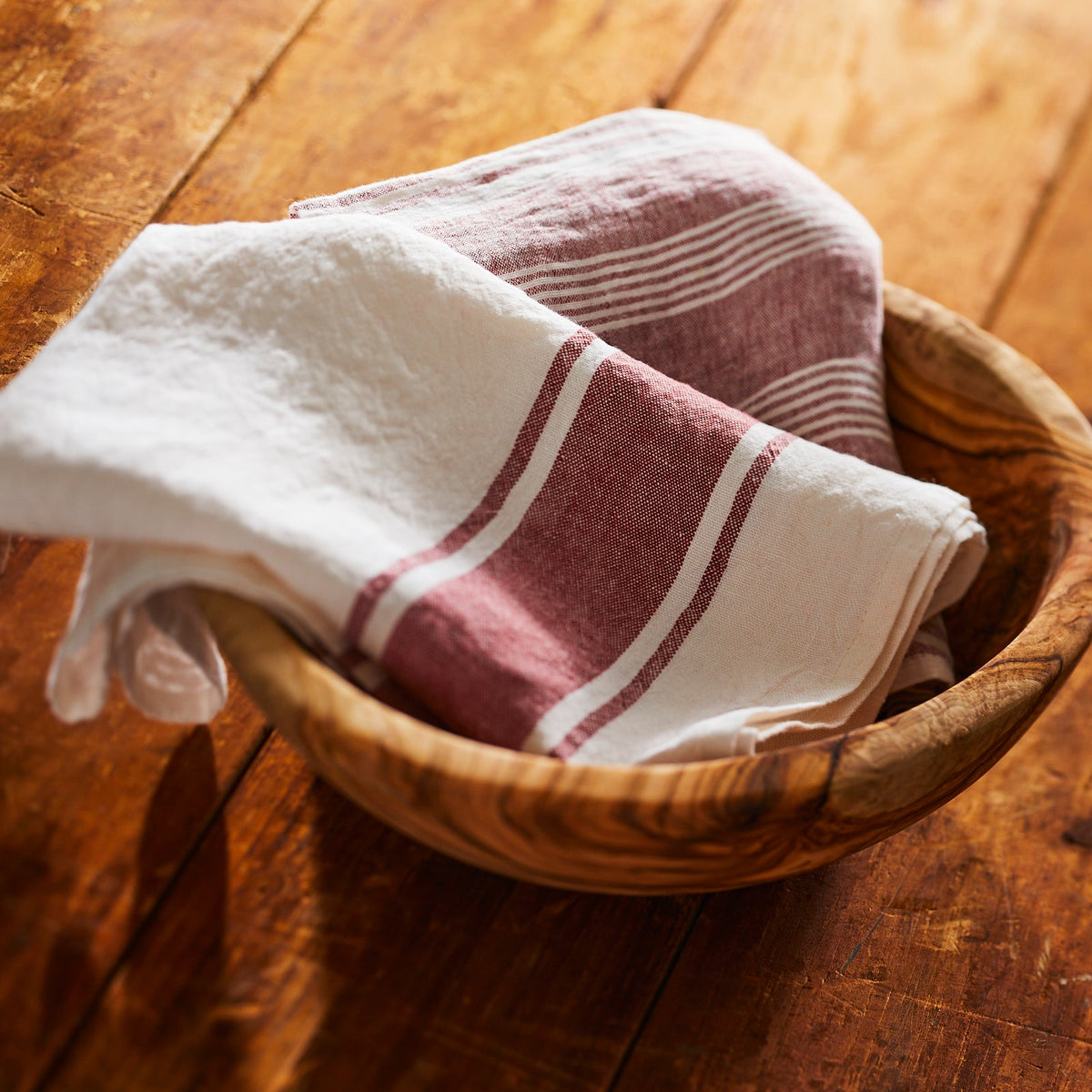Two Trattoria Rosso Linen Kitchen Towels Set/2 in a wooden bowl on top of a woven wooden table.