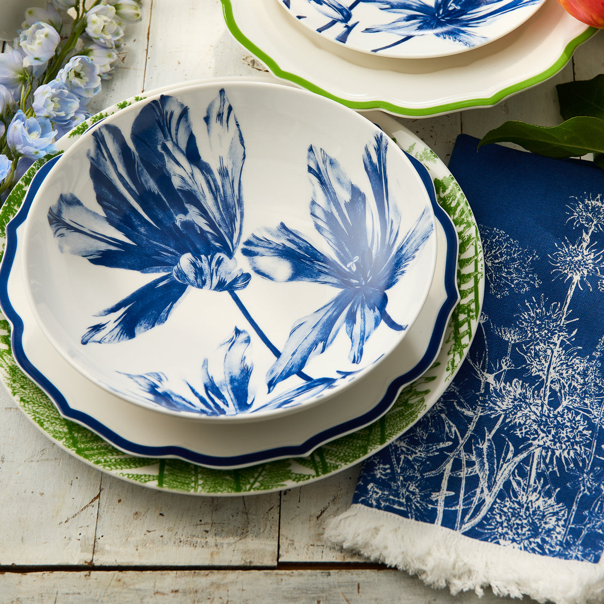 A stack of three floral plates, featuring the Caskata Tulip Entrée Bowl, and a blue napkin with white botanical print arranged on a rustic wooden table.