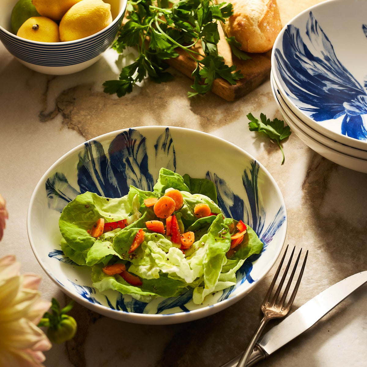 A fresh salad with lettuce, cherry tomatoes, and carrot slices is served in the Caskata Tulip Entrée Bowl, placed on a marble surface alongside stacked plates, cutlery, lemons, parsley, and bread.