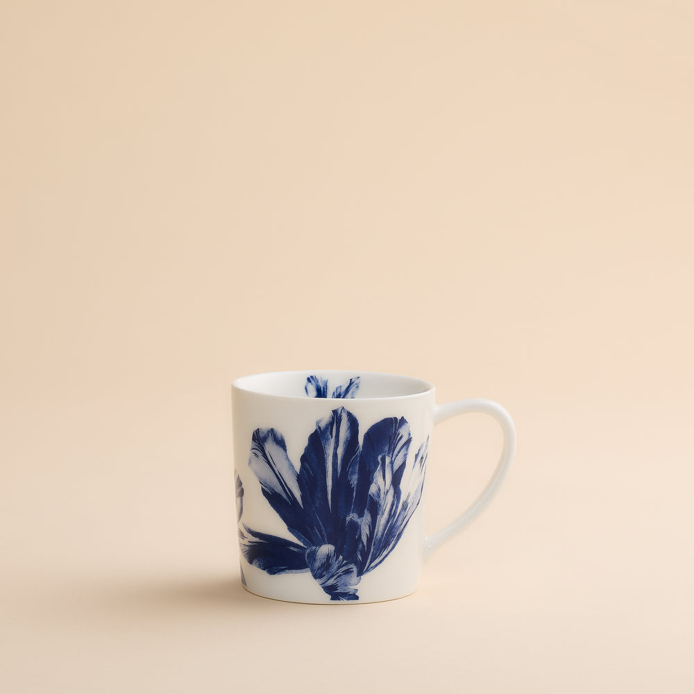 The Caskata Tulip Mug features a delicate blue botanical floral design on white high-fired porcelain, combining elegance and durability. Perfect for your favorite beverages, this mug is shown on a light beige surface with a matching neutral background.