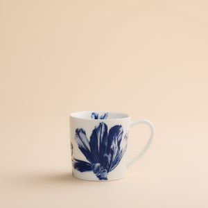 The Caskata Tulip Mug features a delicate blue botanical floral design on white high-fired porcelain, combining elegance and durability. Perfect for your favorite beverages, this mug is shown on a light beige surface with a matching neutral background.