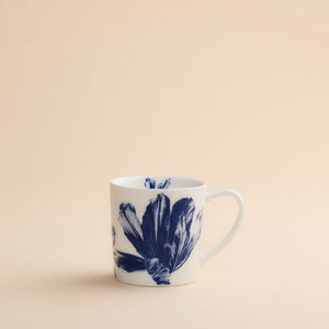 The Caskata Tulip Mug features a delicate blue botanical floral design on white high-fired porcelain, combining elegance and durability. Perfect for your favorite beverages, this mug is shown on a light beige surface with a matching neutral background.