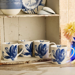 Four Caskata Tulip Mugs in white porcelain with blue floral designs are displayed on a rustic wooden shelf, accompanied by a vase of flowers and ceramic dishes.