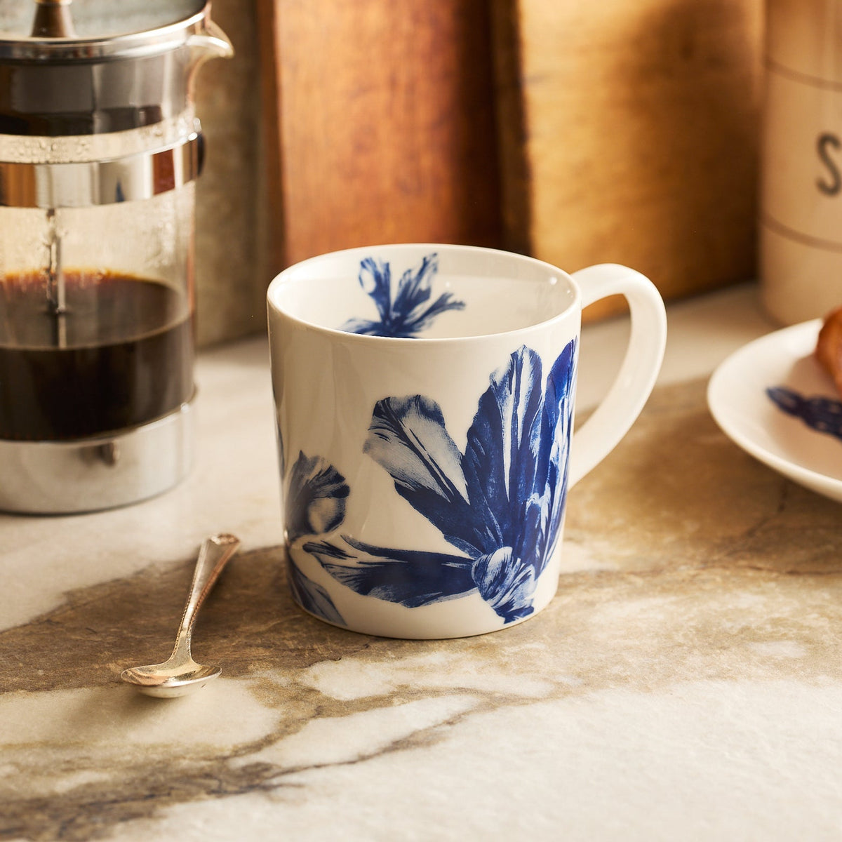 The Caskata Tulip Mug, featuring blue botanical designs, rests on a marble countertop beside a French press, a spoon, and a plate of food.