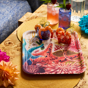 The Caskata Underwater Affair Large Rectangular Birchwood Tray features a coral reef design and holds shrimp cocktails with lime and dill, drinks, and festive paper ornaments on a wooden table.