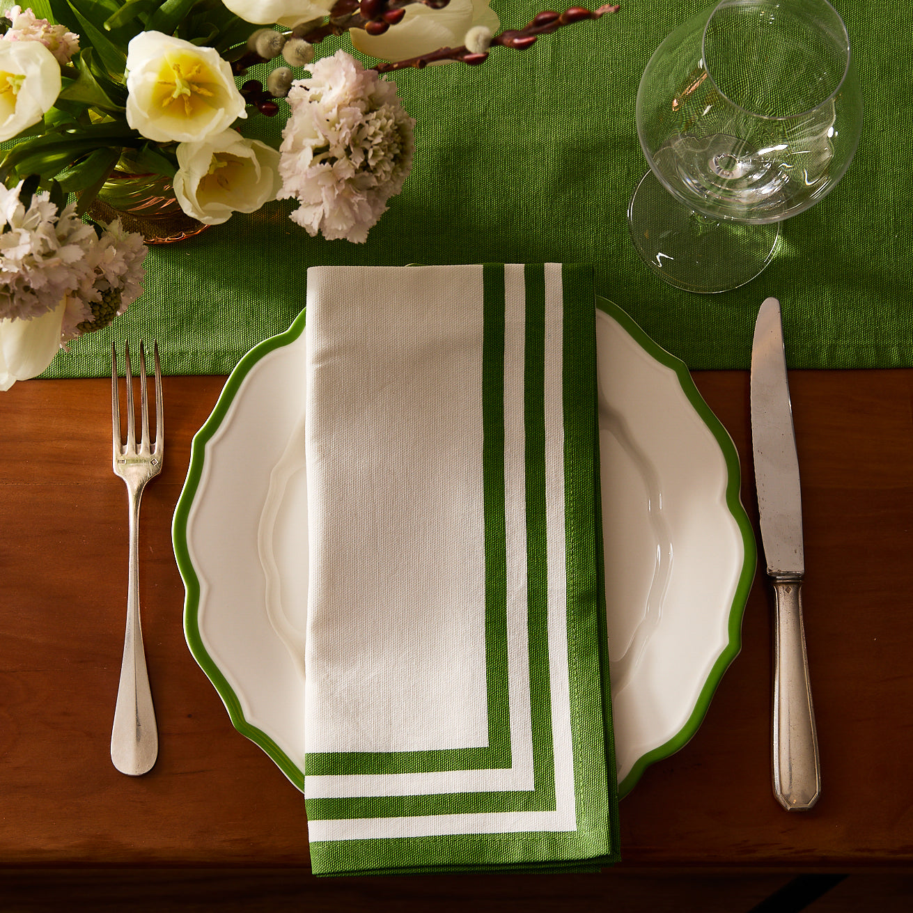 Four white Caskata Triple Stripe Verde Dinner Napkins, crafted from machine washable cotton with parallel green borders, are neatly stacked in a slightly fanned arrangement.