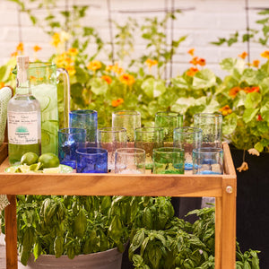 A wooden cart with tequila, a pitcher, and limes sits by potted plants, featuring Caskata’s Viento Medium Blue Tumblers (set of 6) crafted from recycled glass.
