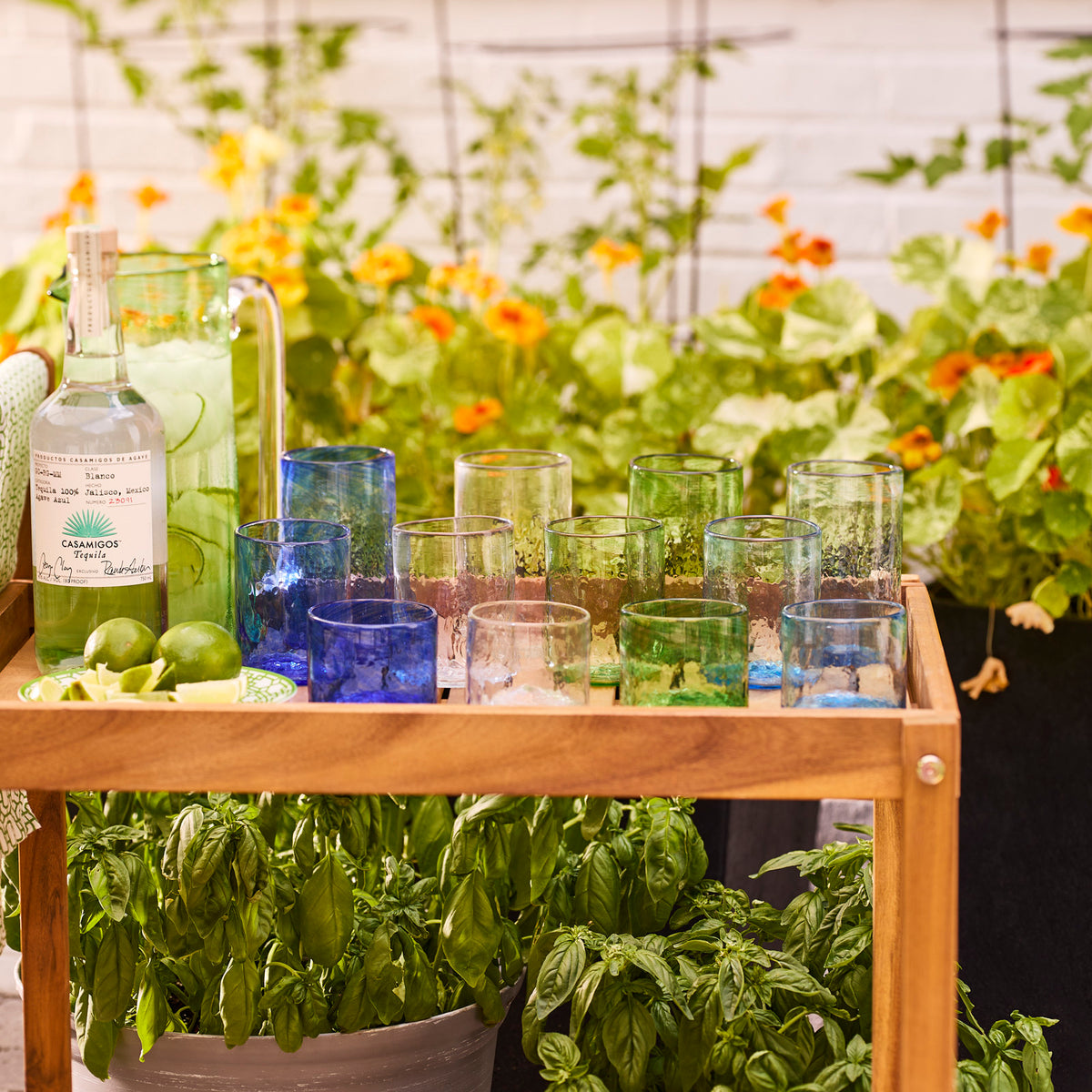 The "Reach for the Sky" Serving Bundle by Caskata features assorted handblown tumblers, a bottle of tequila, a recycled glass pitcher with lime drink, and limes, displayed on a wooden cart among green plants and orange flowers in a garden.