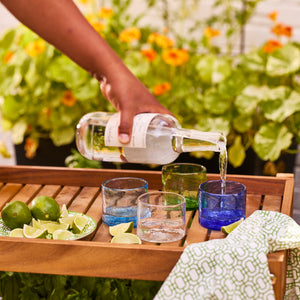 A hand pours a clear liquid into a glass on a wooden tray with limes, lime wedges, and Caskata's Viento Small Cobalt Tumblers (set of 6), crafted from recycled glass. A green-patterned cloth completes the scene.