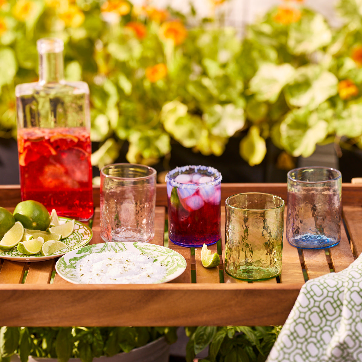A wooden tray with Caskata&#39;s Viento Medium Green Tumblers (set of 6), a jug of red drink, lime wedges, and a plate of salt is arranged outdoors among green plants and orange flowers.