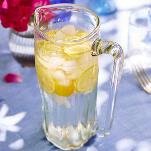 The Caskata Viento Large Clear Pitcher, handblown and filled with ice, lemon slices, and a clear liquid, sits on a blue tablecloth next to a vase of pink flowers.