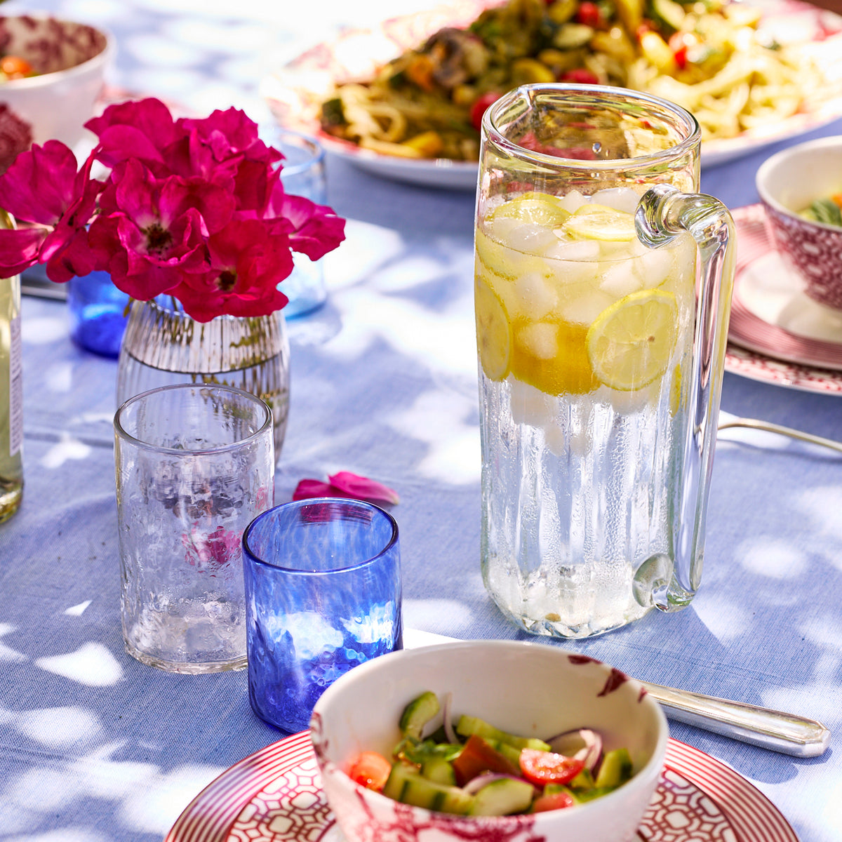 The Caskata Viento Large Clear Pitcher, artisan crafted from recycled glass, holds iced lemon water on a blue tablecloth beside glasses, salad, pasta, and a vase of pink flowers.