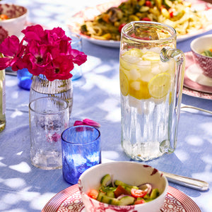 The Caskata Viento Large Clear Pitcher, artisan crafted from recycled glass, holds iced lemon water on a blue tablecloth beside glasses, salad, pasta, and a vase of pink flowers.