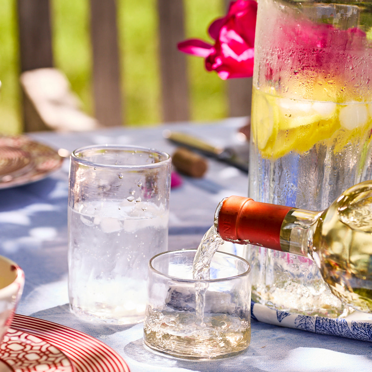 A person pours white wine on a sunlit table set with a pitcher of lemon water, two glasses with ice, red-striped plates, and Caskata Viento Small Clear Tumblers (Set of 6).