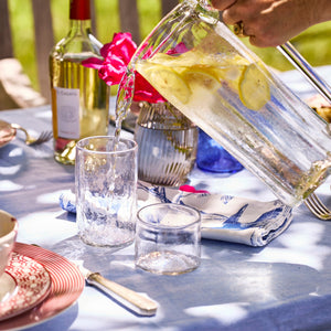 A hand pours lemon water from a recycled glass pitcher into a Caskata Viento Large Clear Tumbler from the set of 6, on an outdoor table adorned with dishes, a wine bottle, and flowers.