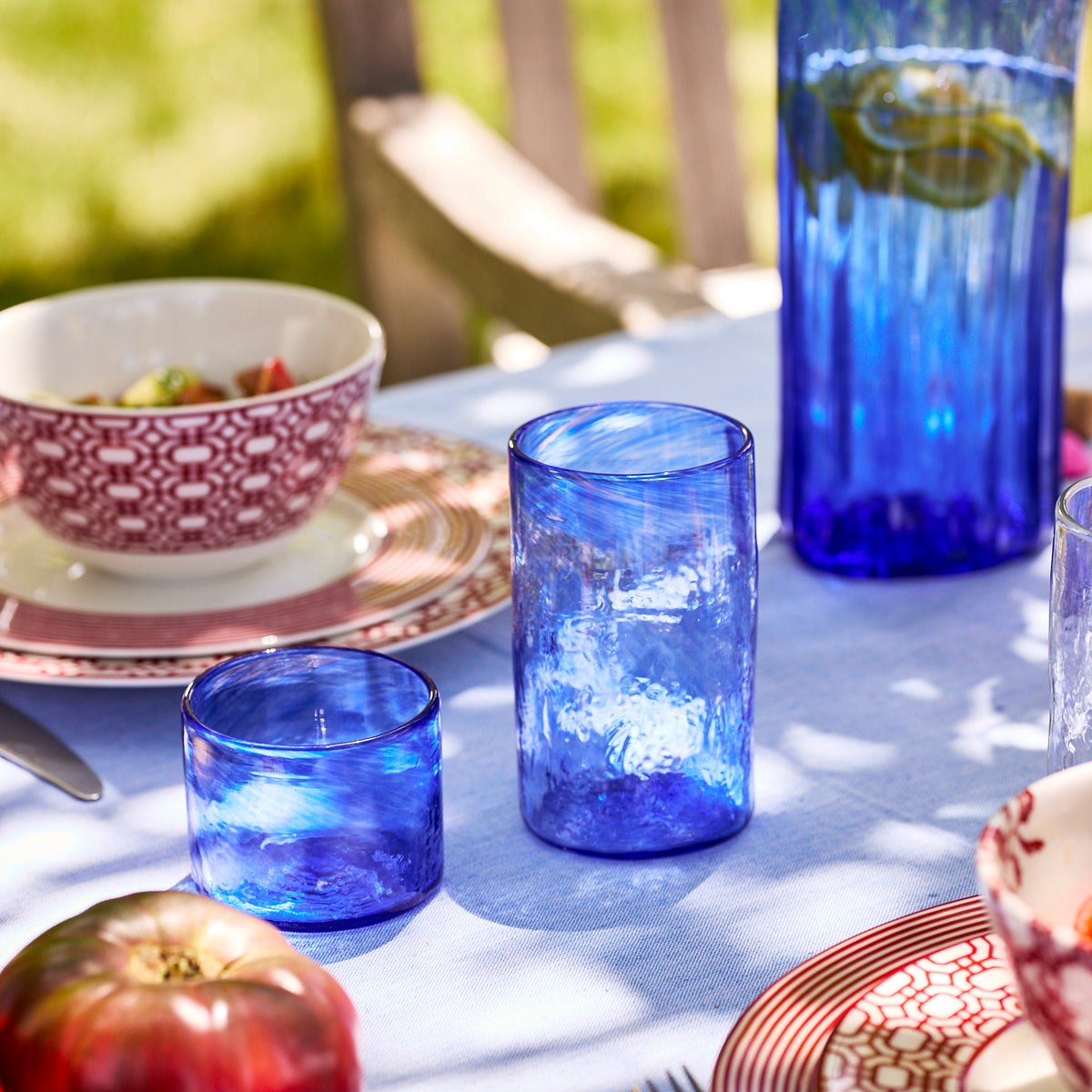 Caskata Viento Small Cobalt Tumblers, Set of 6, made from recycled glass, are displayed on an outdoor table with patterned dishes, tomatoes, and a bowl of salad.