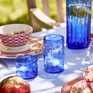 Caskata Viento Small Cobalt Tumblers, Set of 6, made from recycled glass, are displayed on an outdoor table with patterned dishes, tomatoes, and a bowl of salad.