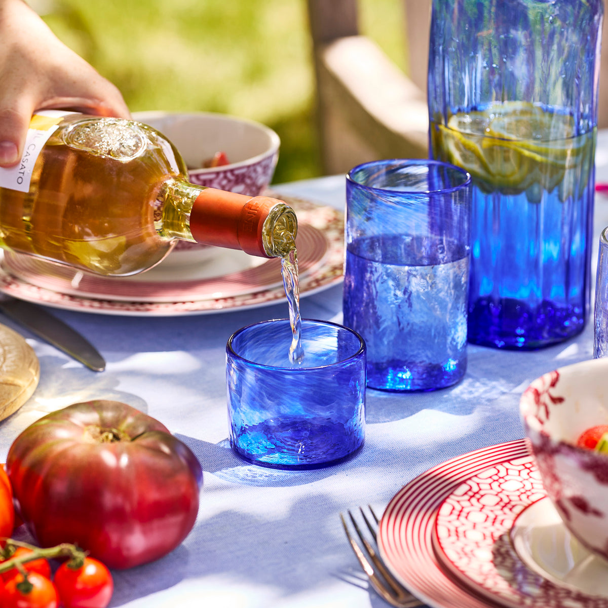 A hand pours white wine into a Caskata Viento Small Cobalt Tumbler from a set of 6 on an outdoor table with patterned plates, recycled glass tumblers, a tomato, and a pitcher with lemon slices.