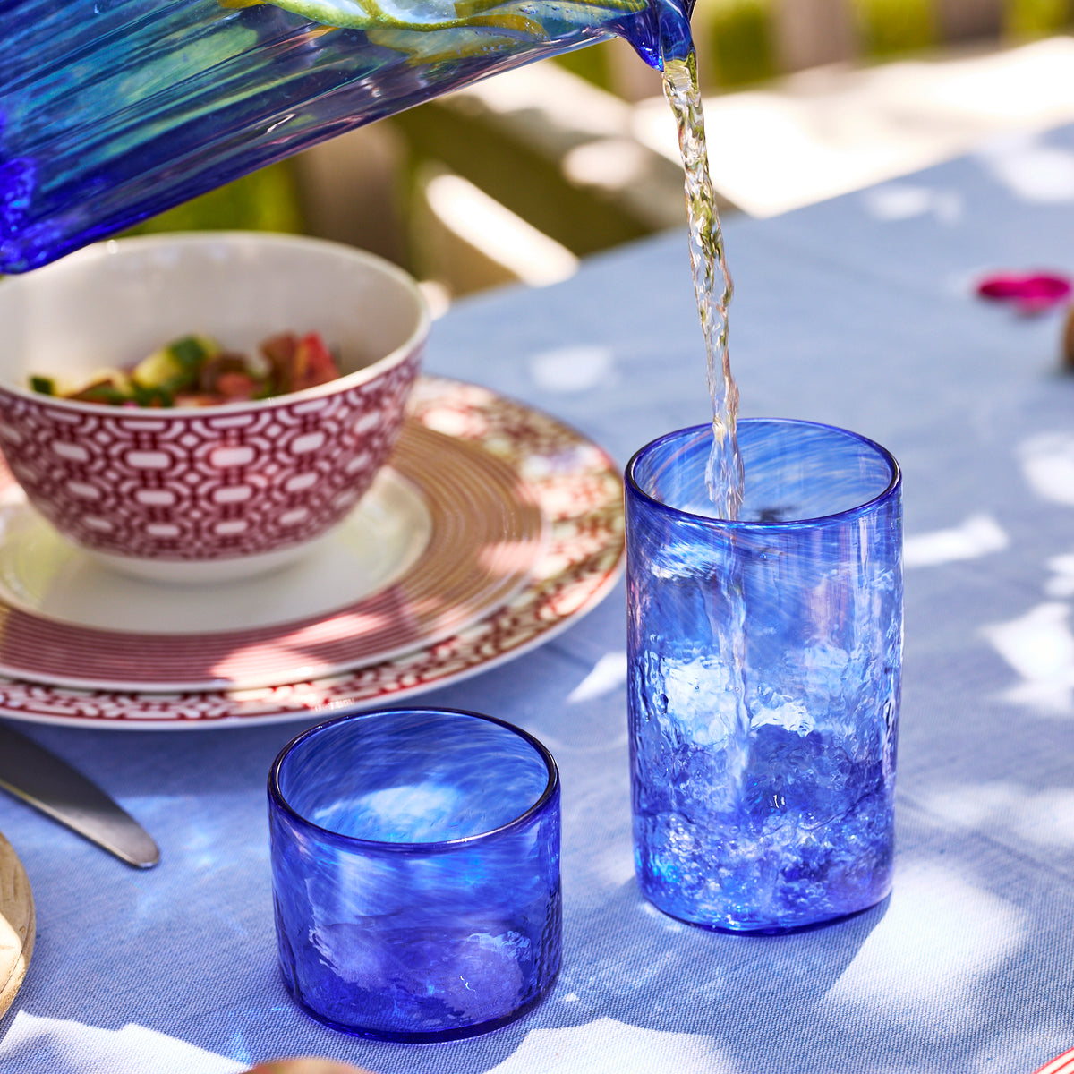 The Caskata "Feeling the Blues" Serving Bundle features a handblown glass pitcher pouring into a cobalt blue tumbler on a table with a bowl and patterned plates—ideal for your coastal bar cart.