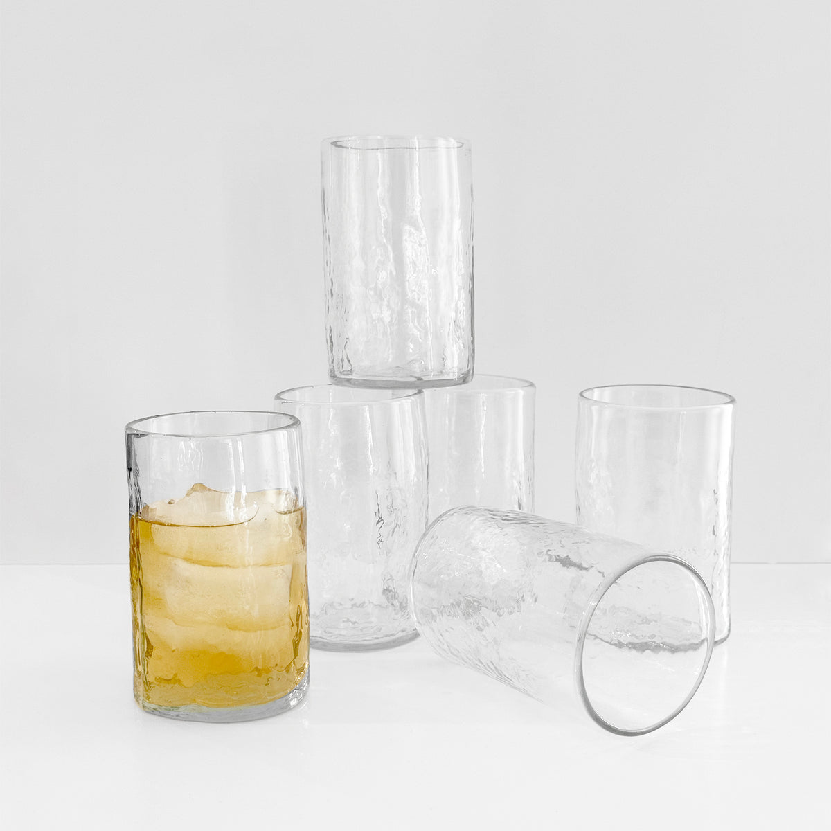 Five clear textured tumblers from the Caskata "Viento Essentials" Serving Bundle are stacked in a pyramid; the front recycled glass tumbler is filled with a yellow iced drink, while the remaining glasses are empty.