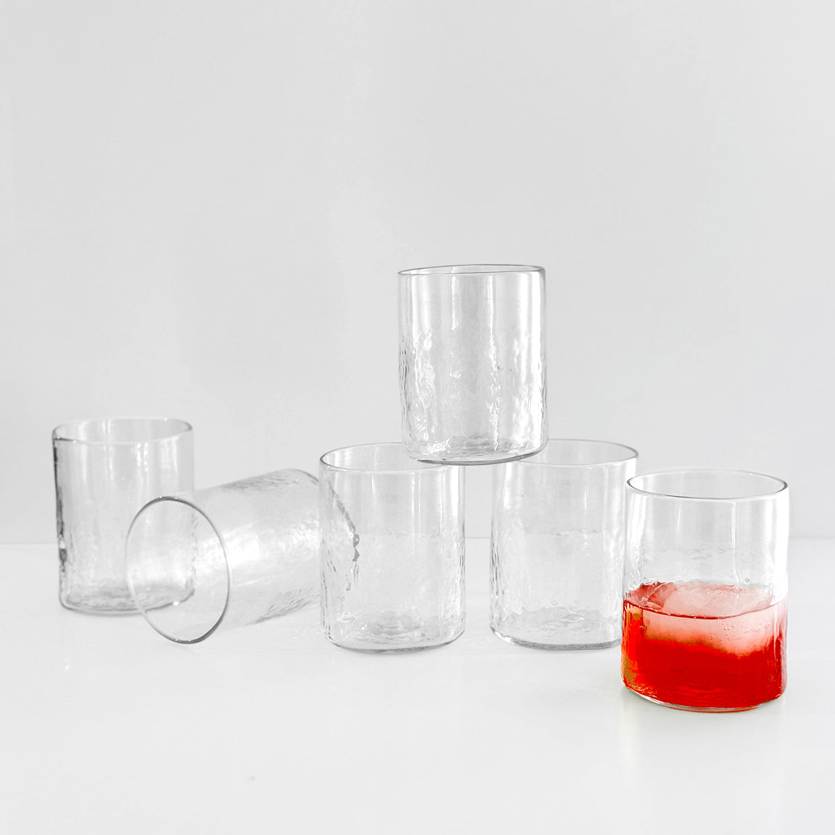Six Viento Medium Clear Tumblers by Caskata are displayed on a white surface—five empty, one holding red liquid with ice—highlighting the artisanal beauty of this handblown recycled glass set.