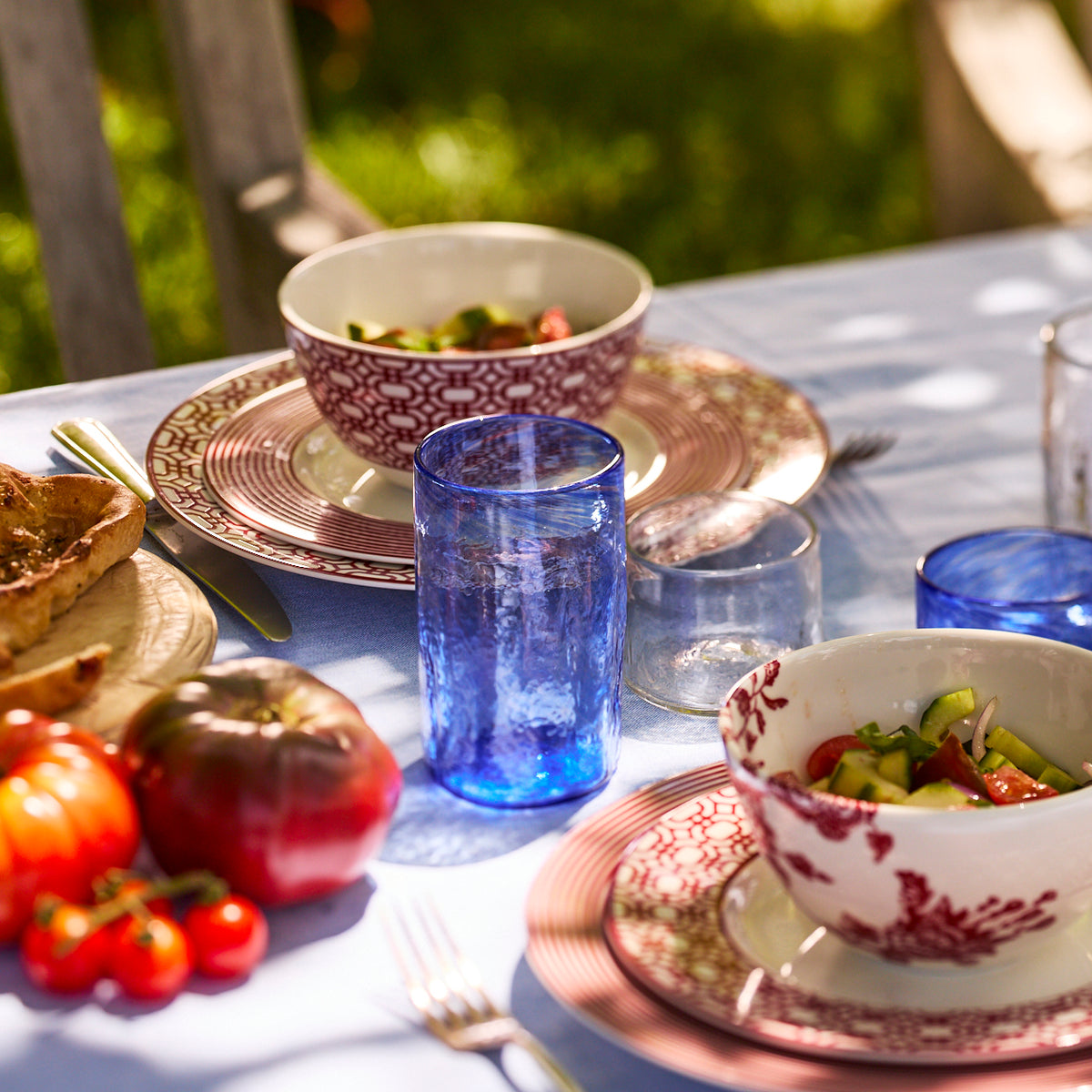 A table set outdoors features patterned plates, Viento Collection salad bowls, fresh tomatoes, bread, and a set of six Caskata Viento Large Cobalt Tumblers on a light blue tablecloth.